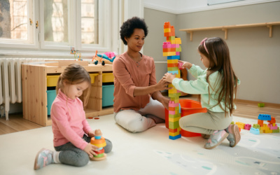 African American teacher and little girls stacking toy blocks while playing on the floor at kindergarten.